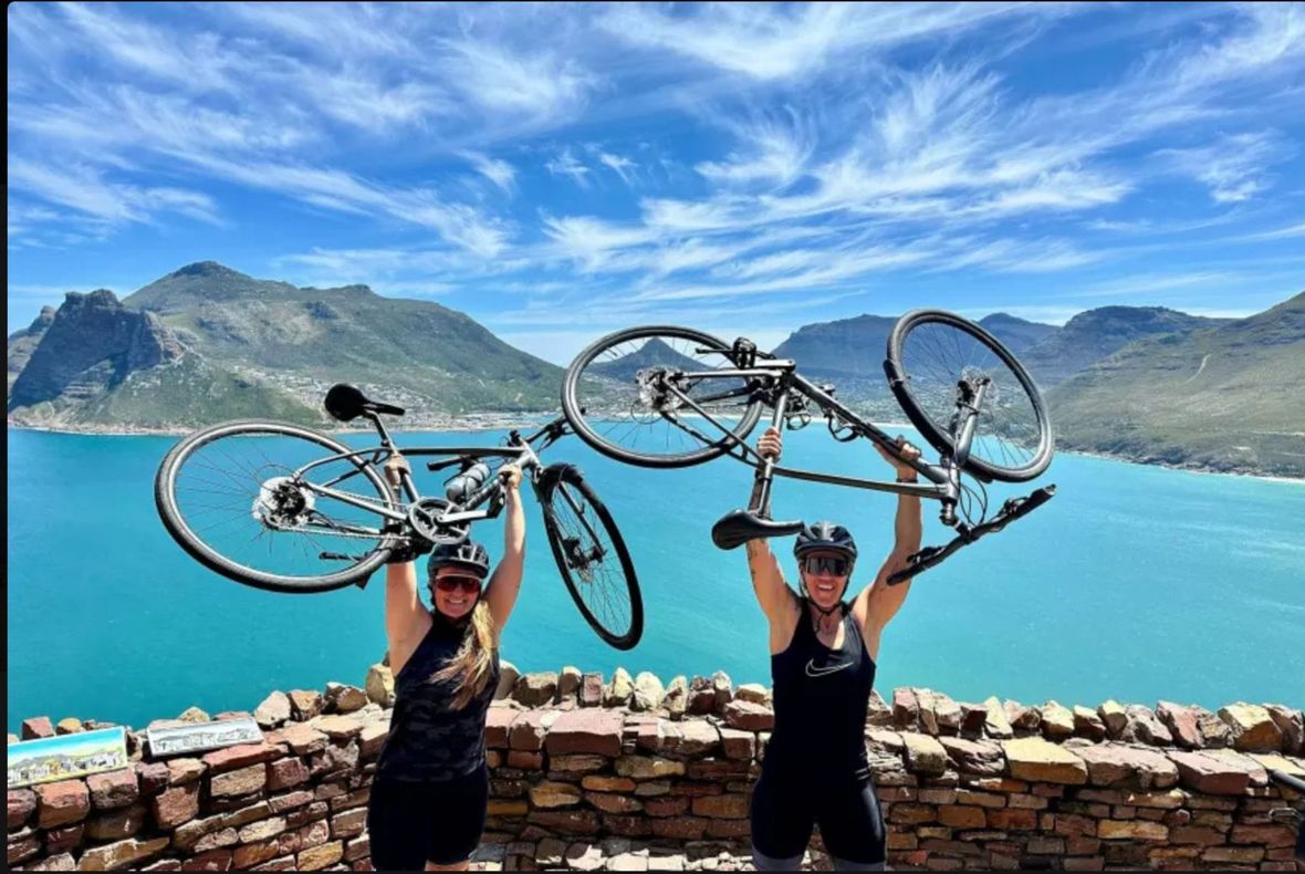 Two female cyclists raising bikes in the air above their heads with a water and mountain backdrop