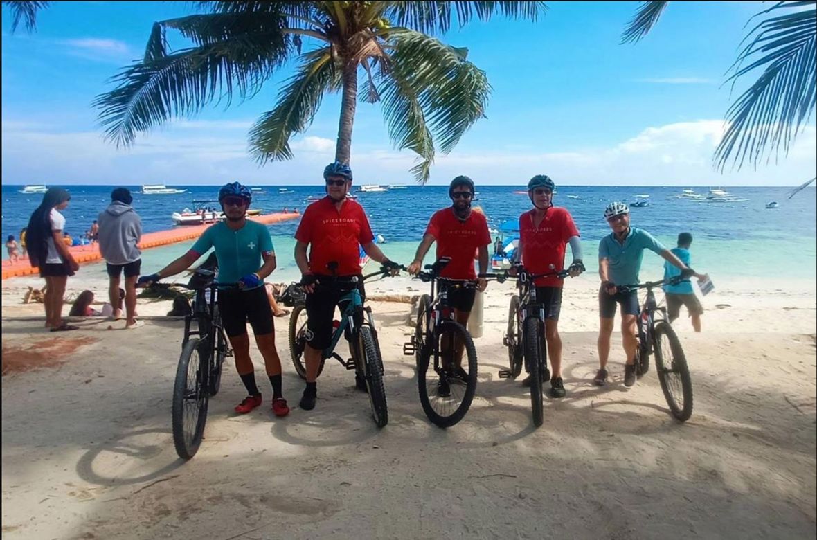 Five people holding their bikes on beach with palm trees.
