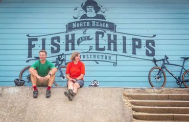 Couple off the bikes, sitting by Fish and Chips large sign