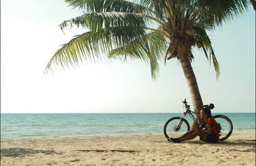 Cyclist sitting on beach by palm tree with bike