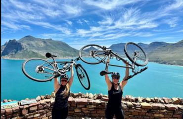 Two cyclists with bikes above head and sea and mountain backdrop