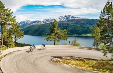 Riders on curved road overlooking lake and mountain