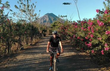 Cyclist riding down bush-lined road with volcano in background