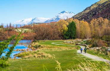 Cyclists on path with greenery to the water then trees and snowy mountains in the distance