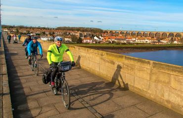 Riders cycling over bridge with town and viaduct behind