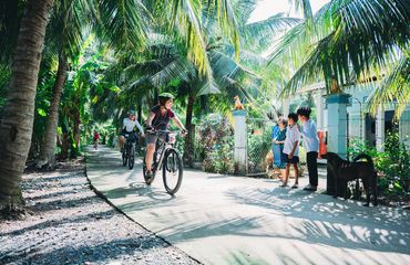 Cyclists riding along palm fringed path with family waving and dog watching