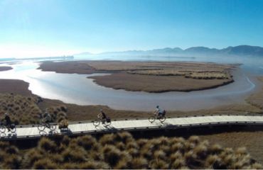 Cyclists riding across a boardwalk with sea and mountain views